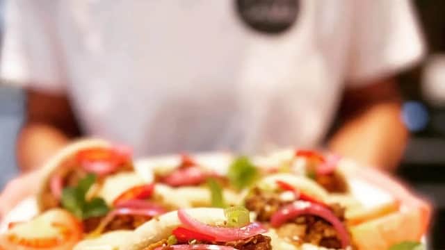 A plate of bao buns filled with shredded meat, garnished with pickled onions, lime wedges, and fresh herbs.