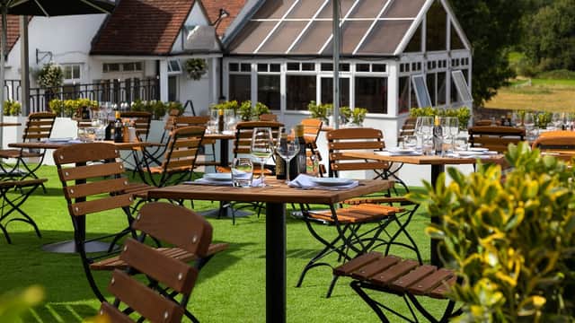 Outdoor dining area with wooden tables and chairs, surrounded by greenery and a glass conservatory in the background.