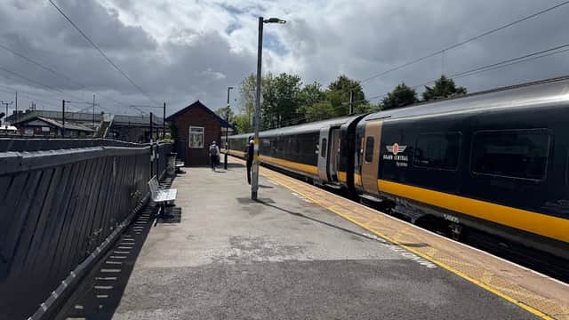 Carlton Miniott Railway Station platform with a train, a passenger, and a station building under a partly cloudy sky.