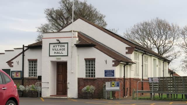 White building with a sign reading "Ayton Village Hall," surrounded by trees and a parking area.