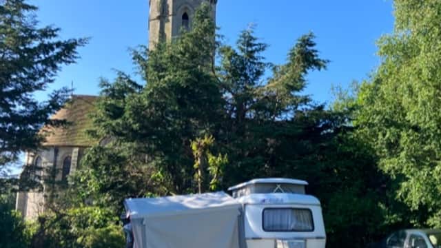 Caravan site with a camper, chairs, and a church tower in the background, surrounded by trees and blue sky.