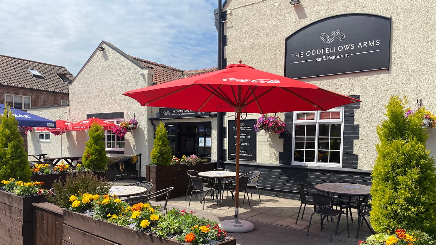Quaint bar entrance with red umbrellas, outdoor seating, and colorful flowers in planters at The Oddfellows Arms, Yorkshire.