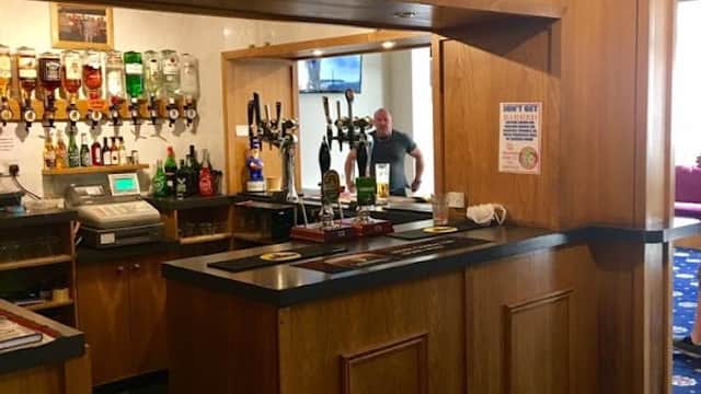 Wooden bar with drinks, a TV on the wall, and a person in the background at Rawcliffe Bridge Club.
