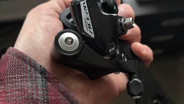 A person holding a black bicycle derailleur with a silver logo, set against a blurred background.