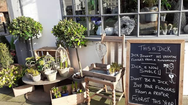 Charming florist display with potted plants, wooden crates, and a humorous chalkboard sign about Valentine's flowers.