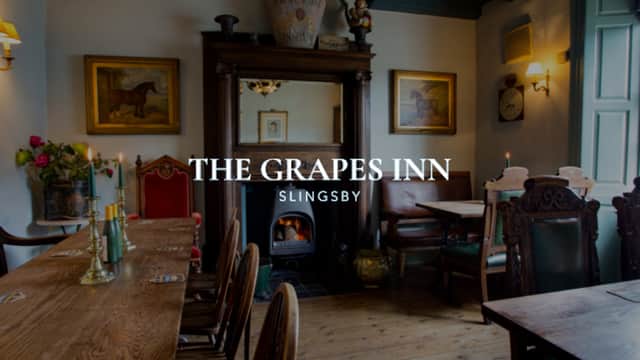 Cozy interior of The Grapes Inn with wooden tables, chairs, a fireplace, and framed artwork on the walls.
