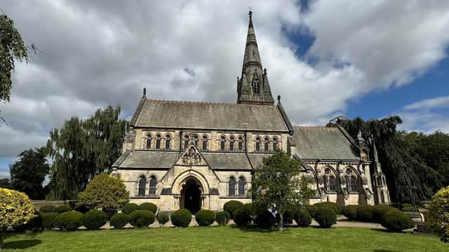 Victorian church with a tall spire, surrounded by manicured lawns and trees under a partly cloudy sky.