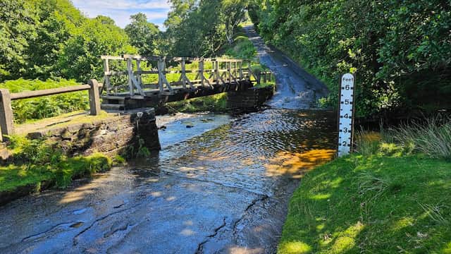 A wooden bridge over a stream with a depth marker, surrounded by lush greenery and a sloping road in Commondale.