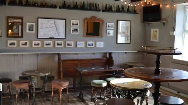 Cozy bar interior with wooden tables, stools, framed photos, and vintage bottles along the walls, illuminated by string lights.