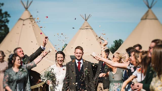 A joyful couple walks through confetti, surrounded by smiling guests and teepee tents at Aldby Park Weddings.