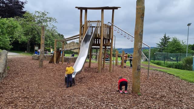 Wooden playground with a slide, climbing structure, and children playing on wood chips, surrounded by greenery.