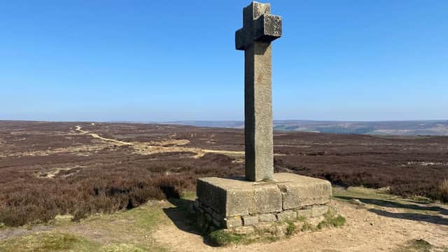 Stone cross on a grassy hilltop with heather-covered landscape and clear blue sky in Lastingham, Yorkshire.