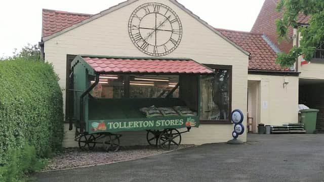 Tollerton Stores features a green cart display, large clock, and a brick building surrounded by greenery in Yorkshire.