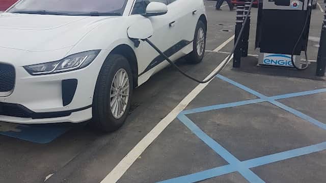 A white electric vehicle charging at a station in West Street Car Park, with buildings visible in the background.