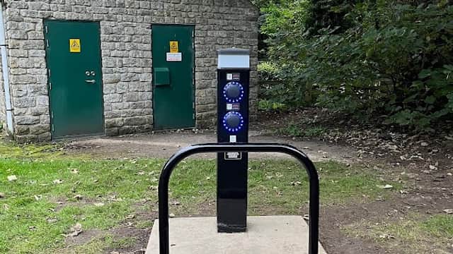 Car park with a charging station in front of a stone building with green doors, surrounded by trees and grass.
