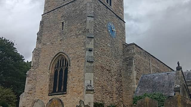 Historic stone church tower with a blue clock, surrounded by gravestones and greenery under a cloudy sky.