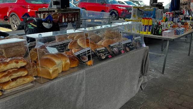 Tables displaying various baked goods and beverages under a wooden structure, with cars parked in the background.