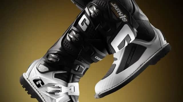 Black and white motocross boots with sleek design, showcasing straps and branding against a gradient background.