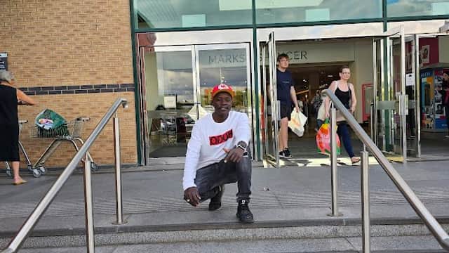 A man in a white sweatshirt kneels on steps outside a shopping center with shoppers entering and exiting.