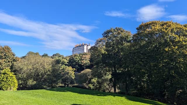 Lush green park with trees and a grand building atop a hill under a bright blue sky with scattered clouds.