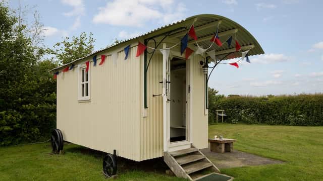 A charming shepherd's hut with a curved roof, bunting, and steps, set in a grassy area with trees in the background.
