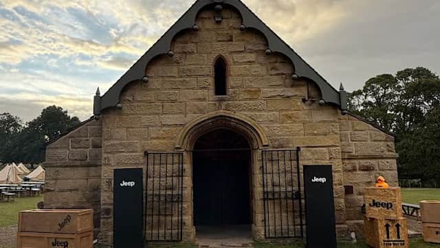 Historic stone building with a pointed roof, flanked by black columns and wooden crates on the lawn under a cloudy sky.