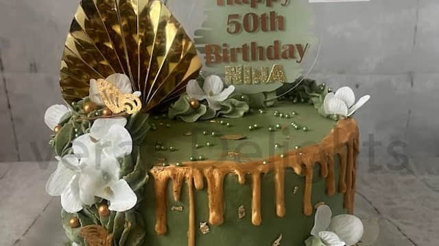 Green birthday cake with gold drip, white flowers, and a "Happy 50th Birthday" topper, adorned with decorative elements.