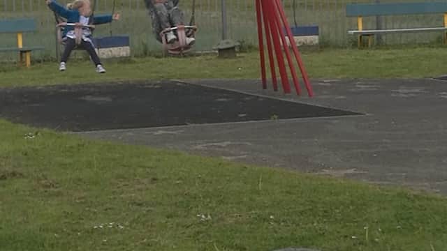 Two children swing on red swings in a grassy park with a blue sky and a building in the background.