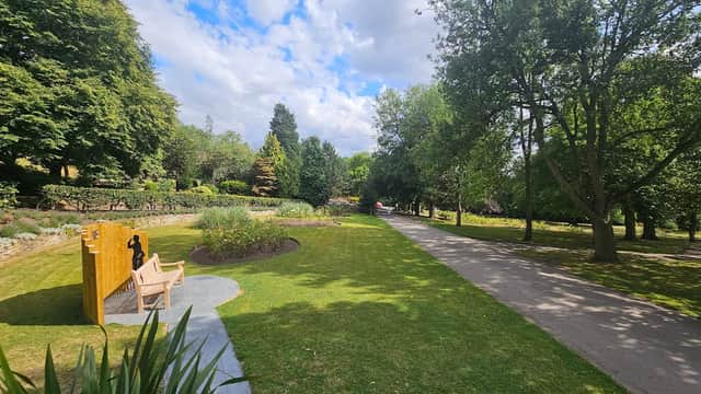 Lush green park with trees, a pathway, and a wooden bench near colorful flowerbeds under a partly cloudy sky.