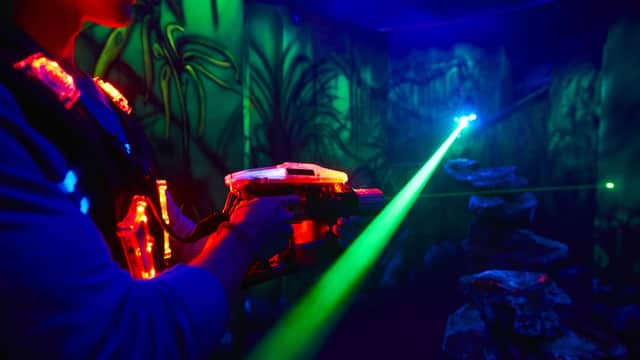 Person playing laser tag in a dark, neon-lit arena with vibrant green and blue lights and jungle-themed walls.