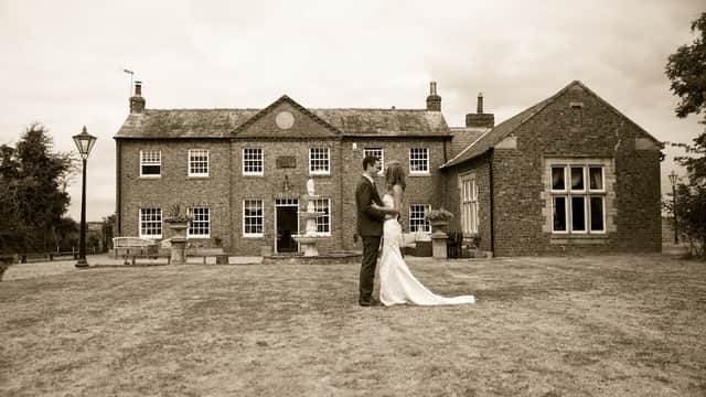 Historic brick building with large windows, a couple in formal attire stands on the lawn, surrounded by greenery.