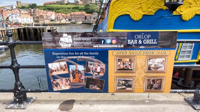 Colorful sign for "The Whitby Endeavour" bar with images and text, overlooking the harbor and buildings of Whitby.