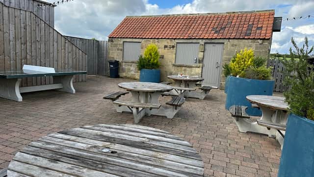 Outdoor seating area with wooden picnic tables, planters, and a rustic building under a cloudy sky.