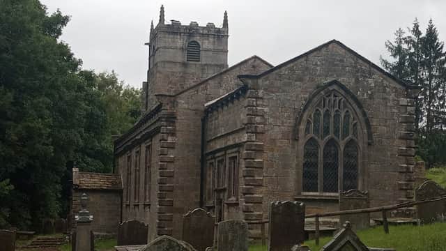 Historic stone church with gothic windows, surrounded by gravestones and lush greenery under a cloudy sky.