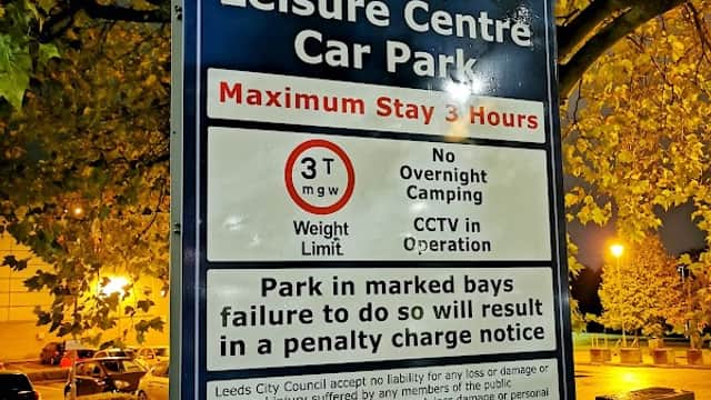 Sign for Morley Leisure Centre Car Park, detailing stay limits, weight restrictions, and CCTV operation. Trees in background.