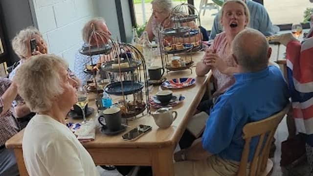 Group of people enjoying afternoon tea at a cafe with large windows, greenery visible outside.