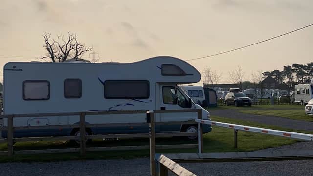 Caravans parked on a grassy site, with trees and a cloudy sky in the background.