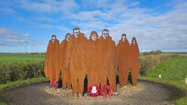 Silhouetted metal figures stand in a circle, surrounded by wreaths, against a blue sky and green fields.