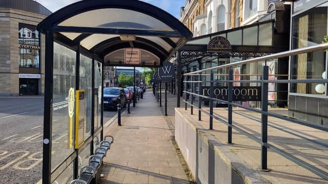 Bus stop shelter with seating, lined pavement, and shops in Harrogate, Yorkshire, under a partly cloudy sky.