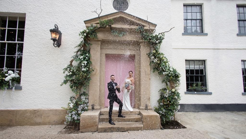 Couple celebrating at The Croft Hotel entrance, adorned with flowers and greenery, with confetti falling around them.