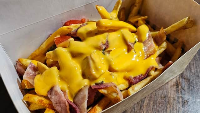 Fries topped with bacon, diced tomatoes, and melted cheese in a cardboard container.