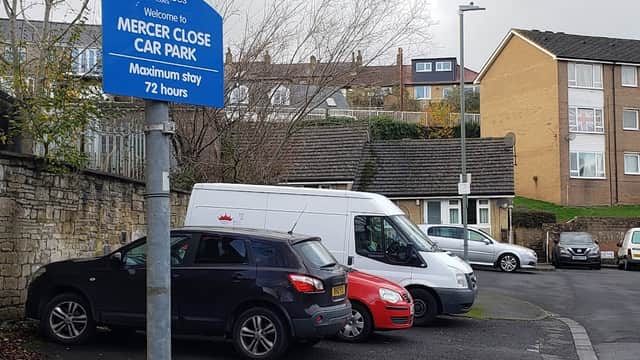 Sign for Mercer Close Car Park with parked cars and residential buildings in the background on a cloudy day.
