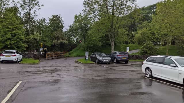 Pod Point Charging Station in a wet, tree-lined parking area with several parked cars and a footpath in the background.