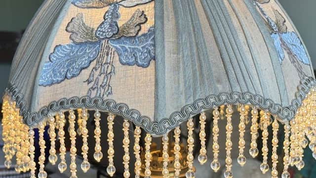 A decorative lamp with a blue floral shade and beaded fringe, resting on a brass base, in a bright interior setting.