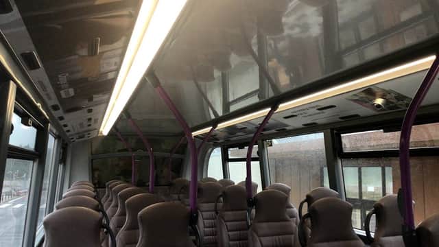 Interior of a bus with purple accents, featuring rows of gray upholstered seats and overhead lighting.