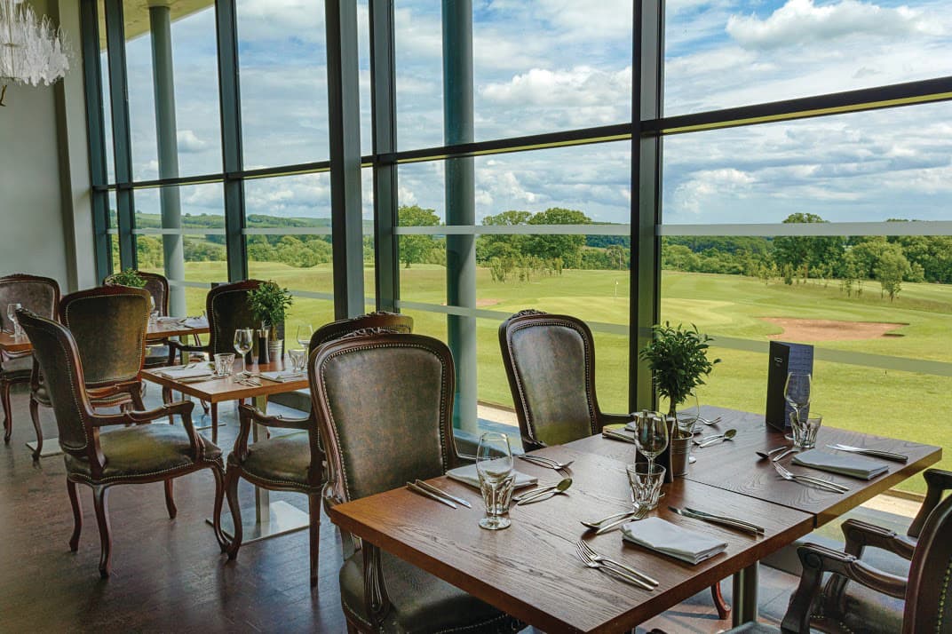 Elegant dining area with wooden tables, plush chairs, and large windows overlooking lush green countryside.