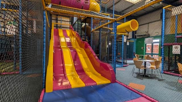 Brightly colored indoor play area featuring a large pink and yellow slide, climbing structures, and seating in the background.
