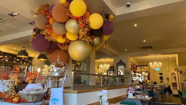 Colorful balloon arrangement hangs above a cozy bakery interior with tables, decor, and patrons enjoying their meals.