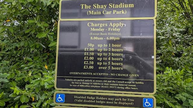 Sign detailing parking charges and conditions at Shay Stadium car park, surrounded by greenery in Exley, Yorkshire.