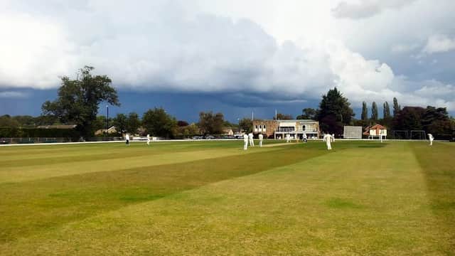 Cricket match in progress on a green field with cloudy skies and trees in the background at Collingham & Linton Cricket Club.